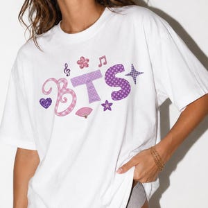 Könnte beinhalten: Ein weißes T-Shirt mit kurzen Ärmeln und dem Schriftzug BTS in großen, violett und rosa gemusterten Buchstaben. Das Design enthält dekorative Elemente wie ein violettes Herz, eine rosa Blume, eine Musiknote, einen kleinen Fächer und einen Stern.