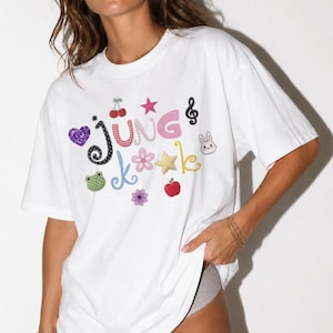May include: A white short-sleeved t-shirt featuring the text JUNG KOOK in colorful, patterned letters. The design includes various icons such as a purple heart, red cherries, a pink star, a treble clef, a white rabbit, a green frog, a flower, a yellow star, and a red apple.
