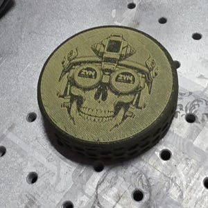 May include: A round, olive-green object with a detailed black graphic of a skull wearing night vision goggles and a helmet. The text "ZVN" is visible on the goggles. The object has a textured edge and appears to be made of wood.