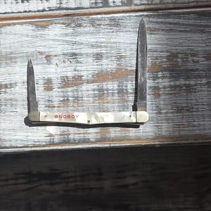 May include: A vintage pocket knife with two blades, one large and one small, both open. The handle is white with the word "SNOBOY" printed on it. The knife is set against a weathered, white-painted wooden surface.