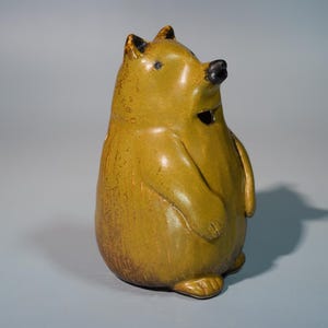May include: A ceramic figurine of a bear with a golden-brown glaze. The bear has a black nose and small, dark eyes. Its ears are pointed, and it has a small, dark opening on its chest. The figurine is standing upright.