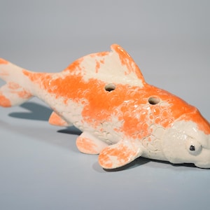 May include: A ceramic koi fish figurine, featuring a white base with orange mottled patterns. The fish has two holes on its back and small black eyes. The figurine is a decorative item.