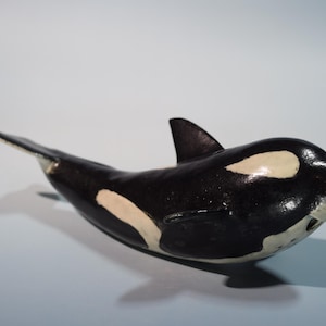 Puede incluir: Figura de orca hecha a mano, predominantemente negra con marcas blancas en el vientre y los ojos. La orca tiene una aleta dorsal y una aleta caudal. La figura está sobre un fondo azul claro.