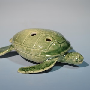 May include: A ceramic turtle figurine in shades of green. The turtle has a textured shell with two holes and outstretched flippers. The figurine is on a light blue surface.