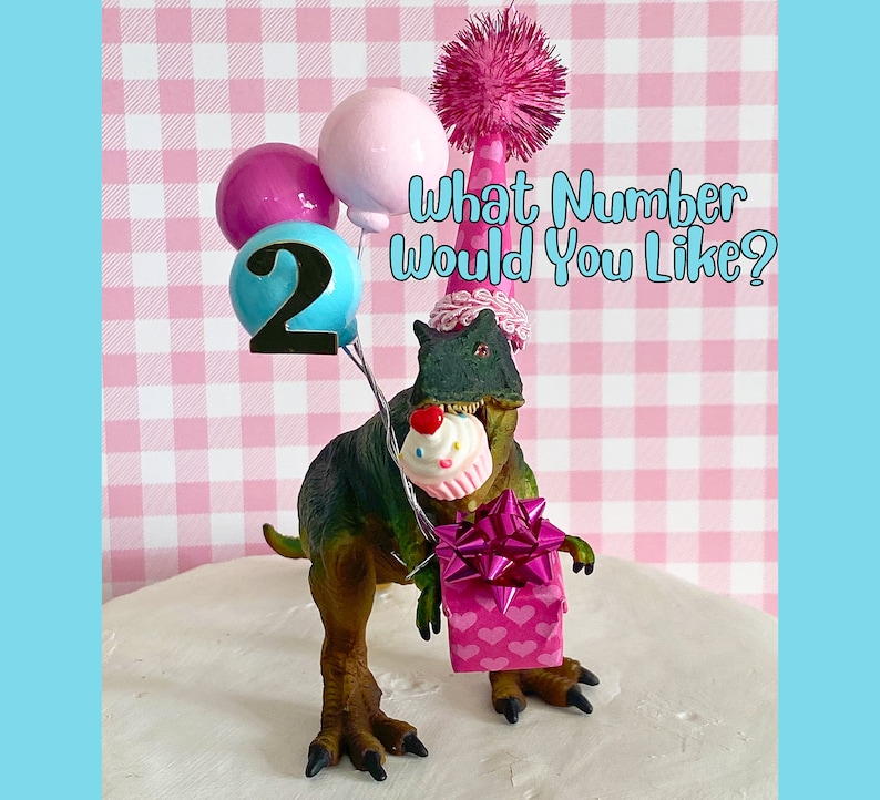 Pode incluir: Um topo de bolo de dinossauro verde usando um chap&eacute;u de festa rosa e branco com um pompom rosa no topo. O dinossauro est&aacute; segurando uma caixa de presente rosa com um la&ccedil;o rosa e um cupcake rosa com uma cereja vermelha no topo. O dinossauro tamb&eacute;m est&aacute; segurando um bal&atilde;o azul com o n&uacute;mero 2 nele. O texto "What Number Would You Like?" est&aacute; escrito em preto sobre um fundo xadrez rosa e branco.