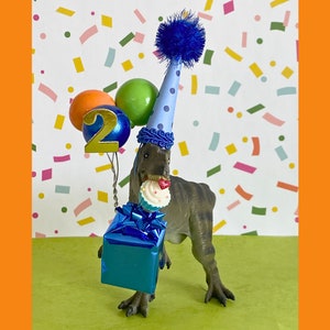 May include: A gray dinosaur figurine wearing a light blue party hat with dark blue polka dots and a blue pom-pom. The dinosaur holds a blue gift box, a cupcake with a red heart, and a cluster of balloons with a gold number two attached.