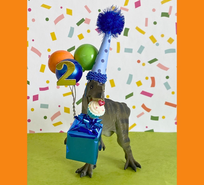 May include: A gray dinosaur figurine wearing a light blue party hat with dark blue polka dots and a blue pom-pom. The dinosaur holds a blue gift box, a cupcake with a red heart, and a cluster of balloons with a gold number two attached.