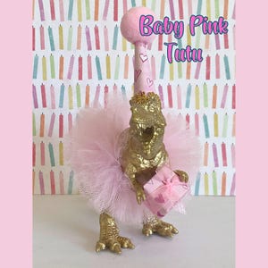 May include: A gold-colored dinosaur figurine wearing a light pink tulle tutu and a matching party hat with a pom-pom top. The dinosaur holds a small pink gift box with a ribbon. The text Baby Pink Tutu is visible above the dinosaur figure.