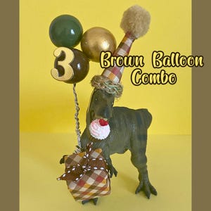 May include: A green dinosaur figurine wearing a plaid party hat with a pom-pom, holding a brown gift box with a ribbon and a wire stem featuring a dark green balloon, a gold balloon, and a dark brown balloon with the gold number 3 attached.