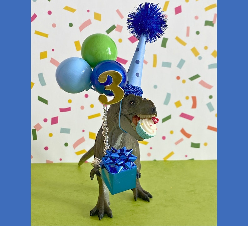 May include: A gray dinosaur figurine wearing a light blue party hat with dark blue polka dots and a blue pom-pom. The dinosaur holds a miniature cupcake in its mouth and a blue gift box with a bow, plus three balloons with a gold number 3.