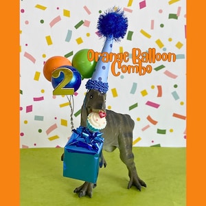 May include: A gray dinosaur figurine wearing a light blue party hat with dark blue polka dots and a blue pom-pom. It holds a blue gift box, a cupcake, and a cluster of balloons with a gold number 2. The text Orange Balloon Combo is visible.