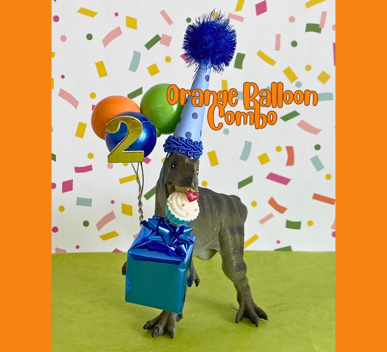 May include: A gray dinosaur figurine wearing a light blue party hat with dark blue polka dots and a blue pom-pom. It holds a blue gift box, a cupcake, and a cluster of balloons with a gold number 2. The text Orange Balloon Combo is visible.