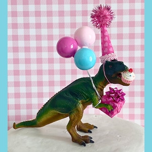 Pode incluir: Um topo de bolo de dinossauro verde usando um chap&eacute;u de festa rosa e branco com bal&otilde;es rosa, azul e branco. O dinossauro est&aacute; segurando um cupcake rosa e branco com um la&ccedil;o rosa.