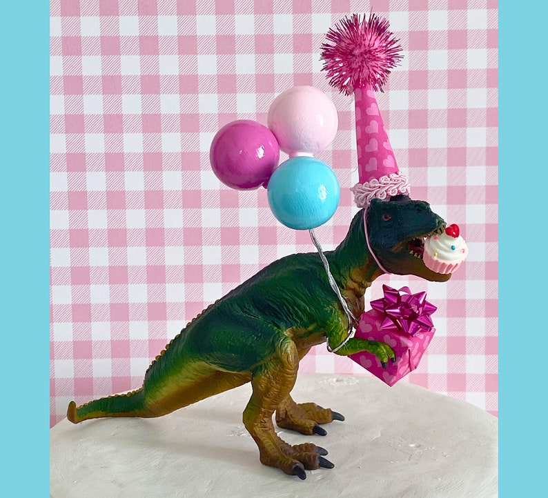 Pode incluir: Um topo de bolo de dinossauro verde usando um chap&eacute;u de festa rosa e branco com bal&otilde;es rosa, azul e branco. O dinossauro est&aacute; segurando um cupcake rosa e branco com um la&ccedil;o rosa.