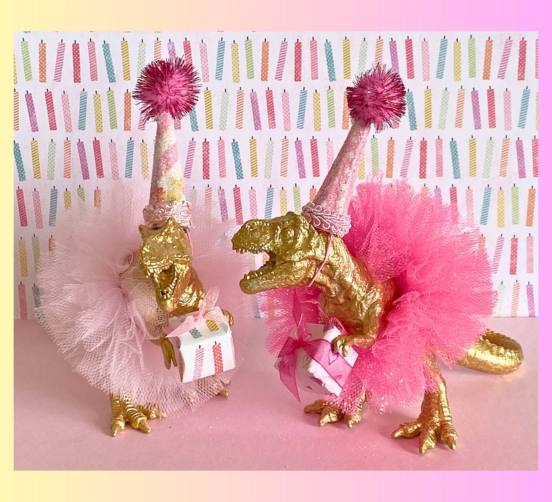 May include: Two gold-colored dinosaur figurines wearing pink party hats with pom-poms and tulle tutus. One dinosaur wears a light pink tutu and holds a small gift box, while the other wears a bright pink tutu and holds a gift box with a pink ribbon.