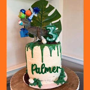 May include: A cylindrical cake with white frosting and green icing drips, featuring a dinosaur figurine wearing a blue party hat, holding a blue gift and a cupcake. A large green leaf, a green number three candle, and the name Palmer are displayed.