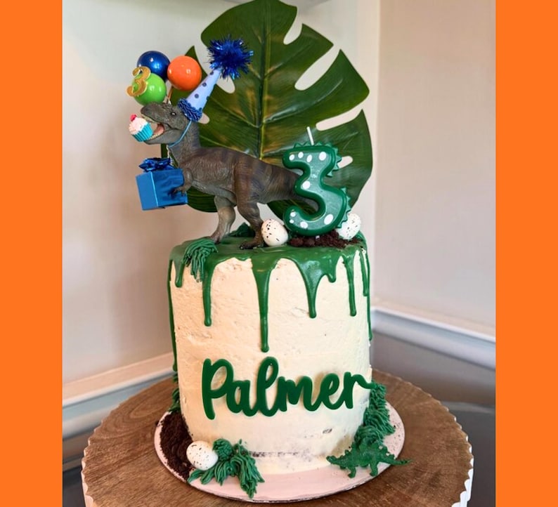 May include: A cylindrical cake with white frosting and green icing drips, featuring a dinosaur figurine wearing a blue party hat, holding a blue gift and a cupcake. A large green leaf, a green number three candle, and the name Palmer are displayed.