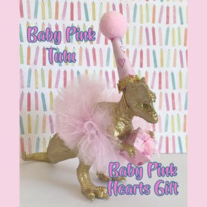May include: A gold-colored dinosaur figurine wearing a light pink tulle tutu and a matching party hat with a pom-pom topper. The dinosaur holds a small pink gift box decorated with heart patterns. Text reads Baby Pink Tutu and Baby Pink Hearts Gift.