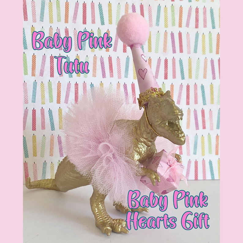 May include: A gold-colored dinosaur figurine wearing a light pink tulle tutu and a matching party hat with a pom-pom topper. The dinosaur holds a small pink gift box decorated with heart patterns. Text reads Baby Pink Tutu and Baby Pink Hearts Gift.
