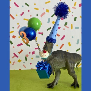 May include: A gray dinosaur figurine wearing a light blue party hat with dark blue polka dots and a blue pom-pom. The dinosaur holds a blue gift box, a miniature cupcake with a red heart, and three balloons, one with the number three attached.
