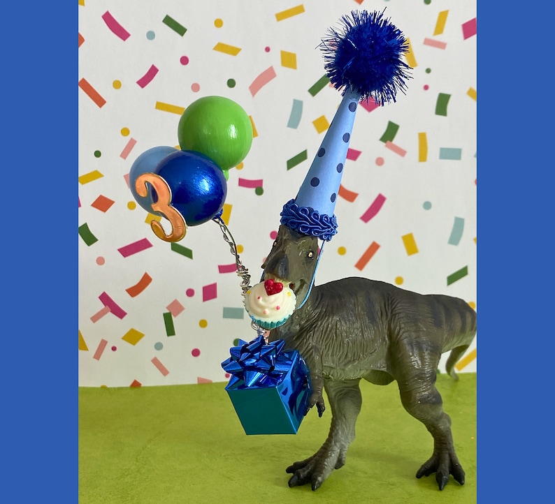 May include: A gray dinosaur figurine wearing a light blue party hat with dark blue polka dots and a blue pom-pom. The dinosaur holds a blue gift box, a miniature cupcake with a red heart, and three balloons, one with the number three attached.