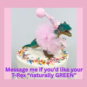 May include: A green T-Rex dinosaur figurine wearing a pink tulle tutu and a matching pink party hat with a pom-pom, standing on a white cake decorated with colorful sprinkles. The text reads: Message me if you'd like your T-Rex naturally GREEN.