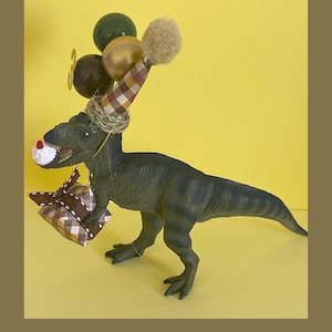 May include: A dark green plastic Tyrannosaurus Rex figurine wearing a brown and white plaid party hat with a tan pom-pom. The dinosaur holds a small gift box wrapped in matching plaid paper and a miniature cupcake in its mouth, with balloons.