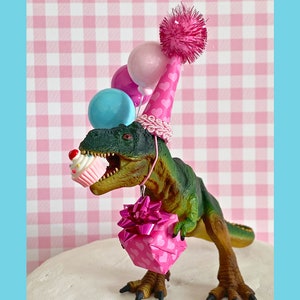 Pode incluir: Um dinossauro verde usando um chap&eacute;u de festa rosa com cora&ccedil;&otilde;es e segurando uma caixa de presente rosa com um la&ccedil;o. O dinossauro tamb&eacute;m est&aacute; segurando um cupcake rosa e branco. H&aacute; bal&otilde;es rosa, azuis e brancos atr&aacute;s do dinossauro.