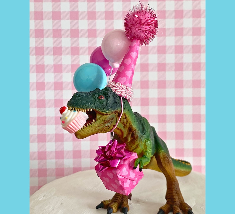 Pode incluir: Um dinossauro verde usando um chap&eacute;u de festa rosa com cora&ccedil;&otilde;es e segurando uma caixa de presente rosa com um la&ccedil;o. O dinossauro tamb&eacute;m est&aacute; segurando um cupcake rosa e branco. H&aacute; bal&otilde;es rosa, azuis e brancos atr&aacute;s do dinossauro.