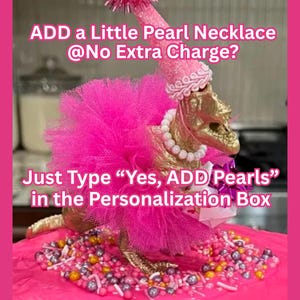 May include: A gold-colored dinosaur figurine wearing a bright pink tulle tutu, a matching pink party hat with a pom-pom, and a white pearl necklace. The text reads: ADD a Little Pearl Necklace @No Extra Charge? Just Type &ldquo;Yes, ADD Pearls&rdquo; in the Personalization Box.