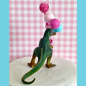 Pode incluir: Topo de bolo de dinossauro verde usando um chap&eacute;u de festa rosa e branco com um pompom rosa no topo. O dinossauro est&aacute; segurando tr&ecirc;s bal&otilde;es: rosa, azul e branco.