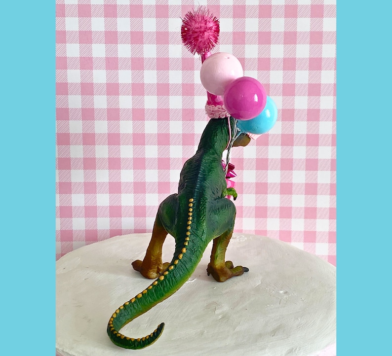 Pode incluir: Topo de bolo de dinossauro verde usando um chap&eacute;u de festa rosa e branco com um pompom rosa no topo. O dinossauro est&aacute; segurando tr&ecirc;s bal&otilde;es: rosa, azul e branco.