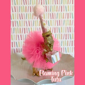 May include: A gold-colored dinosaur figurine wearing a bright pink tulle tutu and a patterned party hat topped with a pink pom-pom. The dinosaur holds a small square gift box decorated with colorful candle illustrations in its front claws.