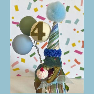 May include: A green dinosaur figurine wearing a striped party hat with a blue pom-pom, holding a cupcake in its mouth. It is decorated with a gold number 4, two small balloons, and a gift box wrapped in striped paper with a sheer ribbon bow.