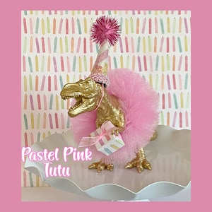 May include: A gold-colored Tyrannosaurus Rex figurine wearing a pastel pink tulle tutu and a matching party hat with a pink pom-pom. The dinosaur holds a small gift box decorated with a candle pattern. The text Pastel Pink Tutu is visible.