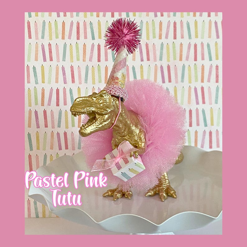 May include: A gold-colored Tyrannosaurus Rex figurine wearing a pastel pink tulle tutu and a matching party hat with a pink pom-pom. The dinosaur holds a small gift box decorated with a candle pattern. The text Pastel Pink Tutu is visible.