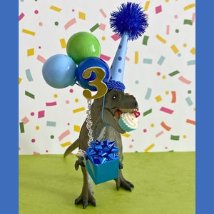 May include: A gray dinosaur figurine wearing a light blue party hat with dark blue polka dots and a blue pom-pom. It holds a cupcake in its mouth, has a gold number three attached, and carries a blue gift box with balloons attached by wire.