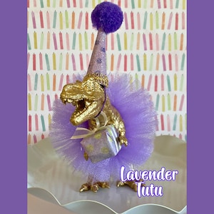May include: A gold-colored Tyrannosaurus Rex figurine wearing a lavender tulle tutu and a matching glittery party hat with a purple pom-pom. The dinosaur holds a small, lavender-patterned gift box tied with a thin, light-colored ribbon.