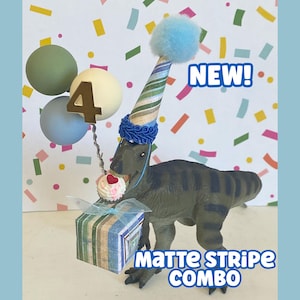 May include: A green dinosaur figurine wearing a striped party hat with a blue pom-pom, holding a striped gift box and a cupcake. A wire holds three spheres and a gold number 4. Text reads NEW! and MATTE STRIPE COMBO. The background features colorful confetti.