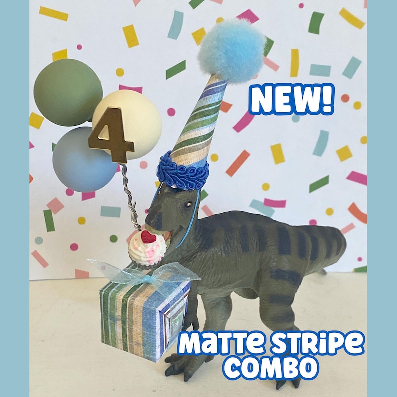 May include: A green dinosaur figurine wearing a striped party hat with a blue pom-pom, holding a striped gift box and a cupcake. A wire holds three spheres and a gold number 4. Text reads NEW! and MATTE STRIPE COMBO. The background features colorful confetti.