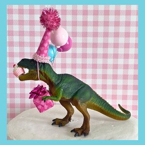 Pode incluir: Um brinquedo de dinossauro verde usando um chap&eacute;u de festa rosa e azul com um pompom rosa no topo. O dinossauro est&aacute; segurando uma caixa de presente rosa e um cupcake rosa e branco. O dinossauro est&aacute; em uma superf&iacute;cie branca com um fundo xadrez rosa e branco.