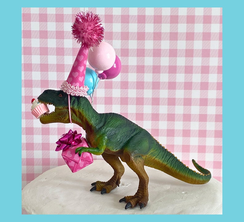 Pode incluir: Um brinquedo de dinossauro verde usando um chap&eacute;u de festa rosa e azul com um pompom rosa no topo. O dinossauro est&aacute; segurando uma caixa de presente rosa e um cupcake rosa e branco. O dinossauro est&aacute; em uma superf&iacute;cie branca com um fundo xadrez rosa e branco.