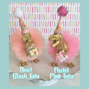 May include: Two gold-colored dinosaur figurines wearing pink tulle tutus and patterned party hats with pom-poms. Each dinosaur holds a small gift box. Text on the image reads New! Blush Tutu and Pastel Pink-Tutu against a candle background.