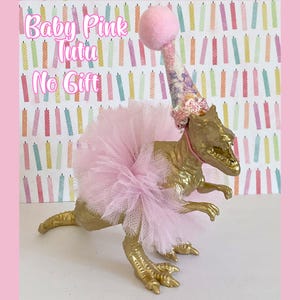 May include: A gold-painted dinosaur figurine wearing a light pink tulle tutu and a matching party hat with a pink pom-pom on top. The text Baby Pink Tutu No Gift is visible in the upper left corner against a background of colorful birthday candles.
