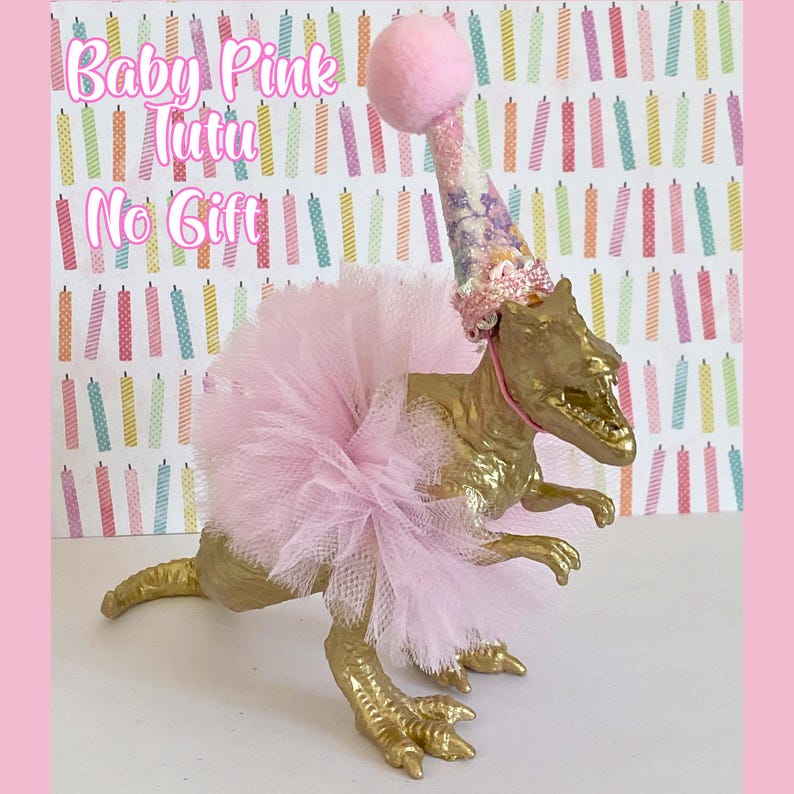 May include: A gold-painted dinosaur figurine wearing a light pink tulle tutu and a matching party hat with a pink pom-pom on top. The text Baby Pink Tutu No Gift is visible in the upper left corner against a background of colorful birthday candles.