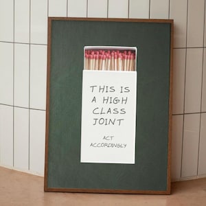 Könnte beinhalten: Gerahmtes Kunstwerk mit einer Streichholzschachtel mit roten Köpfen. Die weiße Schachtel trägt den schwarzen Text "THIS IS A HIGH CLASS JOINT ACT ACCORDINGLY". Der Rahmen ist dunkelbraun, und der Hintergrund ist dunkelgrün.