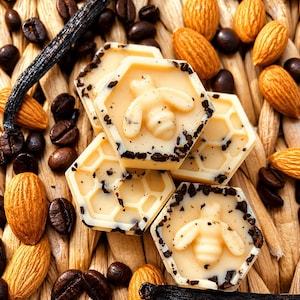 May include: A close-up shot featuring hexagonal sweets with bee motifs, coffee beans, almonds, and a vanilla pod. The sweets are light-coloured with dark flecks. The background has a woven texture. The image evokes a sense of natural ingredients.