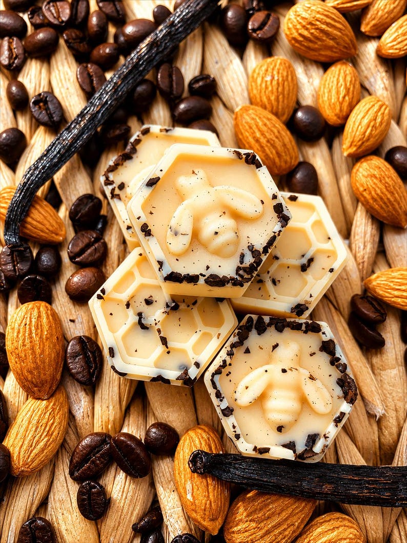 May include: A close-up shot featuring hexagonal sweets with bee motifs, coffee beans, almonds, and a vanilla pod. The sweets are light-coloured with dark flecks. The background has a woven texture. The image evokes a sense of natural ingredients.