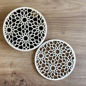 Puede incluir: Dos posavasos redondos de color crema con intrincados patrones florales calados. Los posavasos son de madera y se asientan sobre una superficie de madera. El diseño presenta una serie de formas superpuestas, creando un artículo decorativo y funcional.
