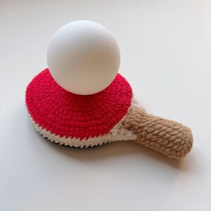 May include: A handmade crochet ping pong paddle with a red head and a tan handle, topped with a white ping pong ball. The paddle has a white border and a black base.
